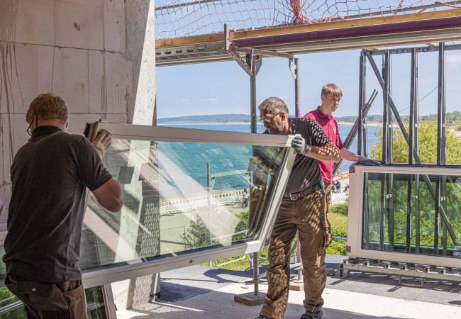 Zwei Männer die in einem Neubau Fenster zur Außenwand tragen